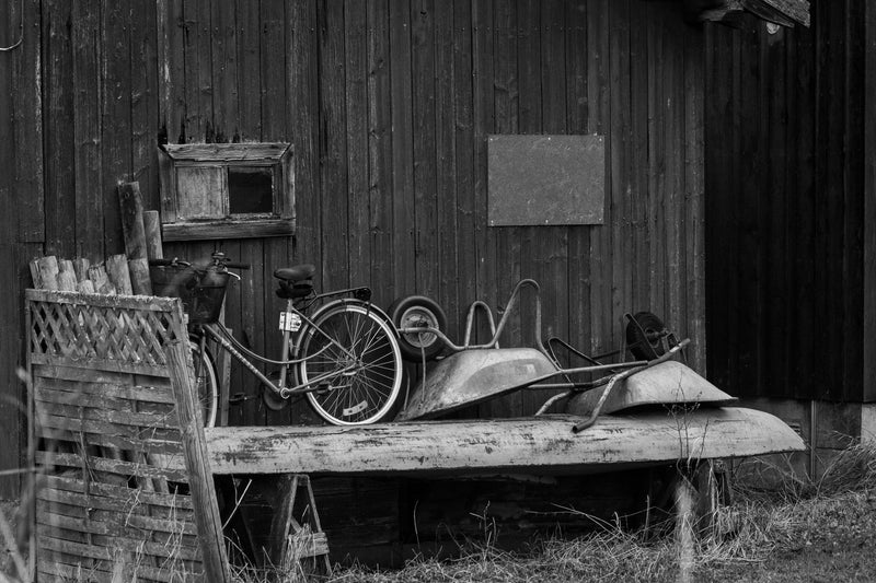 Black and white photo of a vintage bicycle and rusted wheelbarrow beside an old wooden shed.